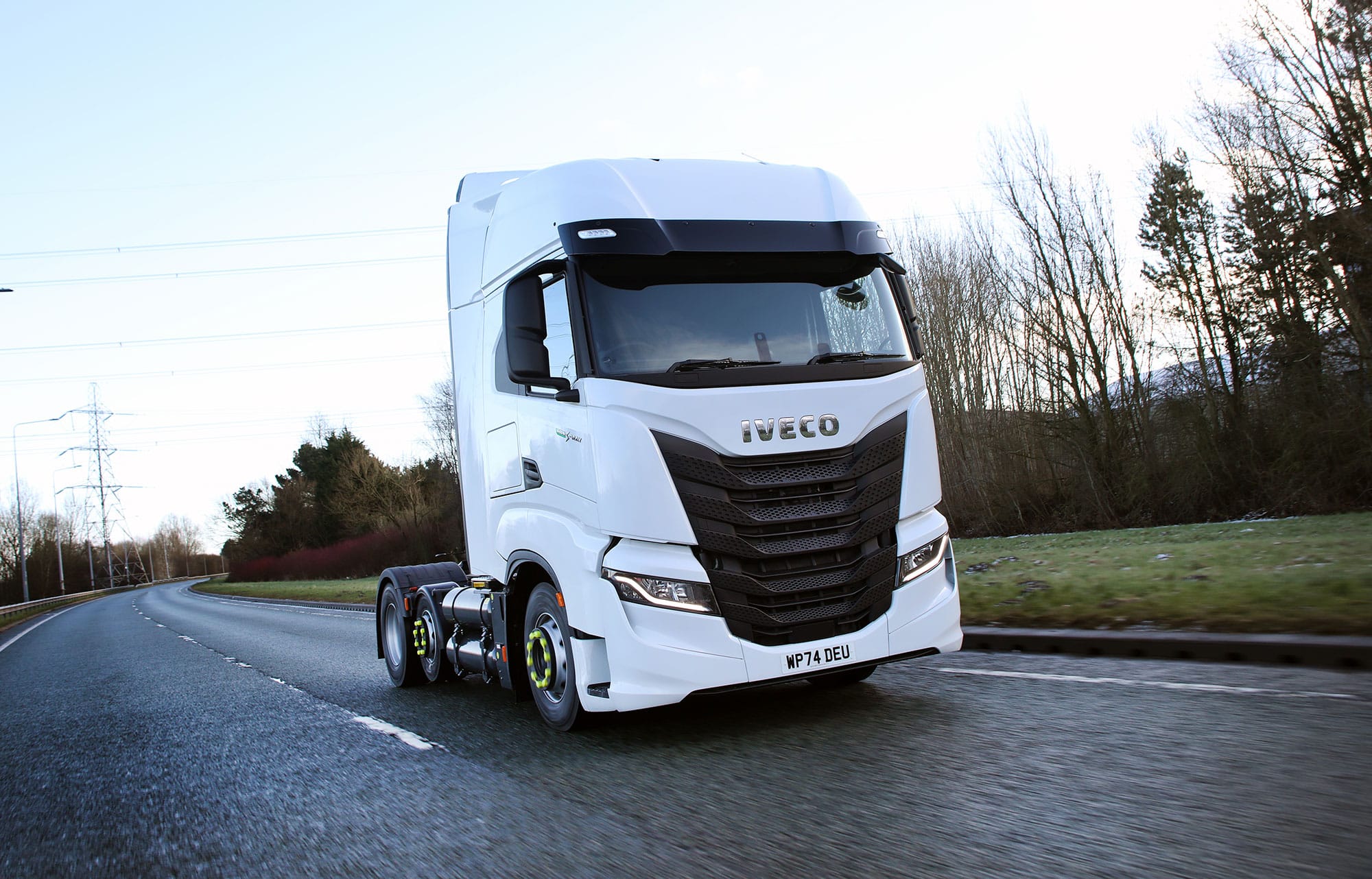A modern white Iveco S-Way CNG 6x2 semi-truck with no trailer test drives on an empty, wet road bordered by grass and bare trees under a cloudy sky after leaving the Astra Vehicle Technologies (AVT) workshop.