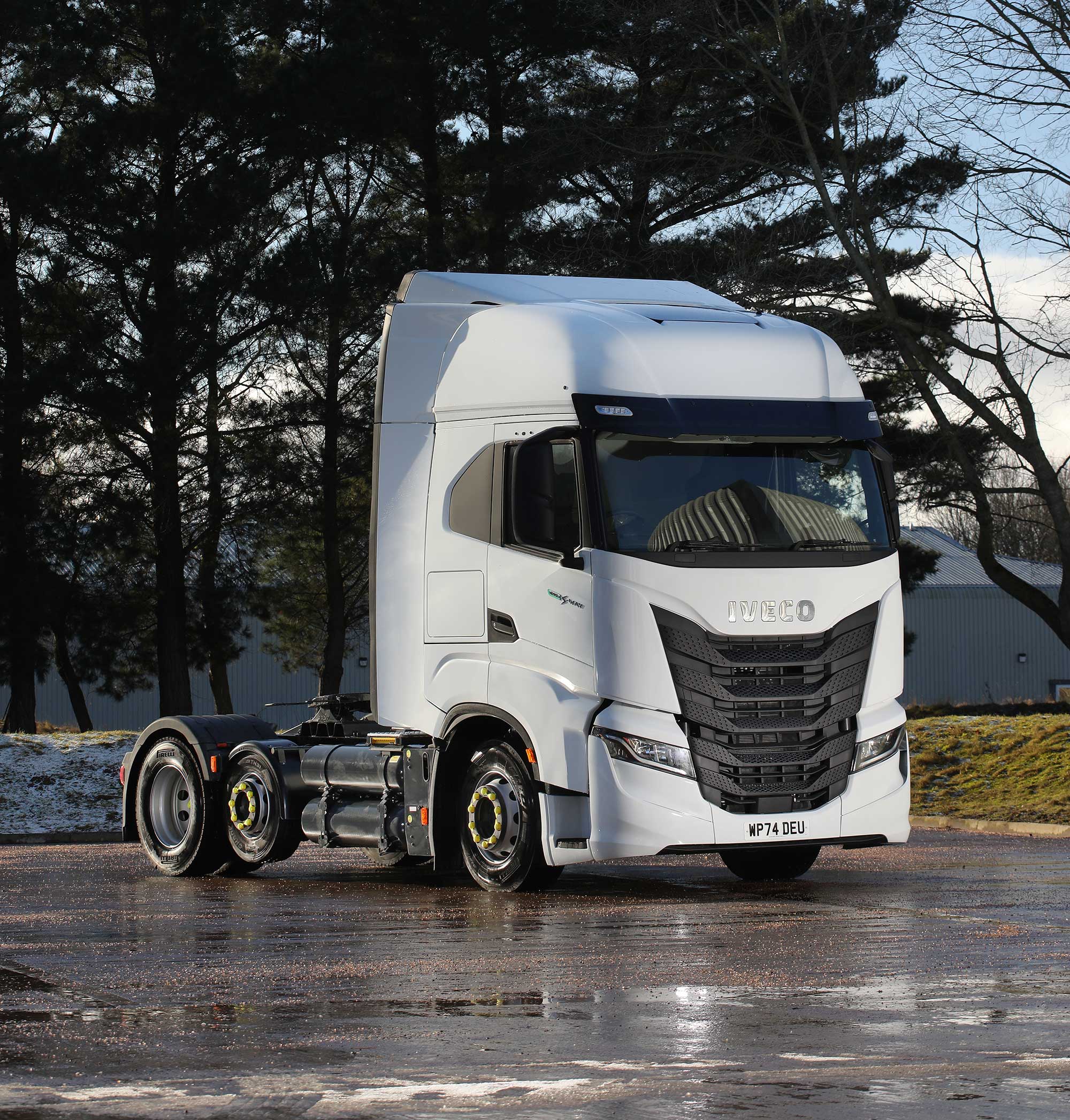 A white Iveco S-Way CNG 6x2 semi-truck cab parked on a wet road with some snow around, surrounded by leafless trees and the Astra Vehicle Technologies (AVT) workshop in the background; sunlight creates reflections on the ground.