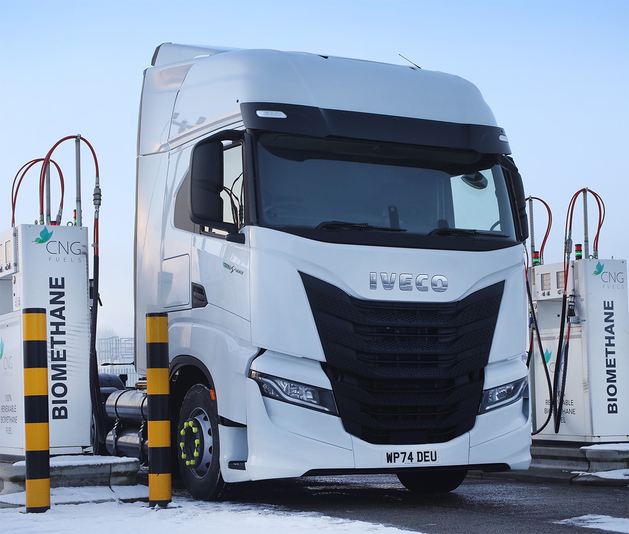 A white IVECO S-Way semi-truck is parked at a biomethane fuel station, refueling at pumps labeled “BIOMETHANE” and “CNG FUEL.” Snow is on the ground, and the sky is clear.