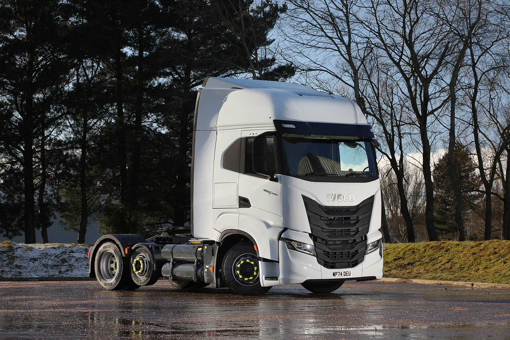 A white Iveco S-Way CNG 6x2 semi-truck cab parked on a wet road with some snow around, surrounded by leafless trees and the Astra Vehicle Technologies (AVT) workshop in the background; sunlight creates reflections on the ground.